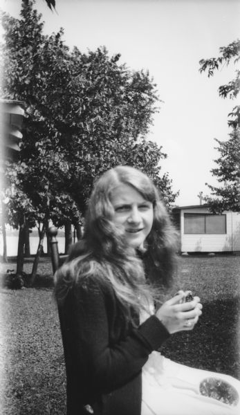 A young woman with long, loose hair sits in a chair in a yard. There is a cottage and lake in the background. She appears to have a mirror in her lap.
