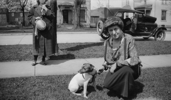 A woman kneels down next to Max, the family dog. Another woman stands on the sidewalk holding a hat, and a car can be seen in the background parked on the street.