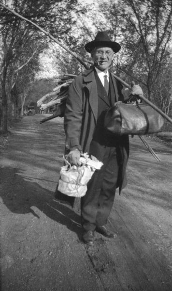 E.W. Brandel, standing in a road. Known as Gramps by Mary Brandel, he is posing holding a bag, a bundle of sticks on his back, and a long stick over his shoulder. He wears eyeglasses and appears to have a pipe or cigarette holder in his mouth.