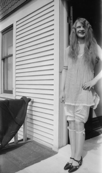 Mary Brandel poses in a doorway on a porch, wearing undergarments, stockings, and shoes. Her hair is loose.
