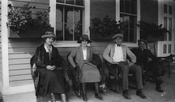 E.W. Brandel, Mrs. Brandel, and the Taylors pose in chairs in front of a building. The man on the right is smoking a cigar, and behind them are windows with flowers in window boxes.