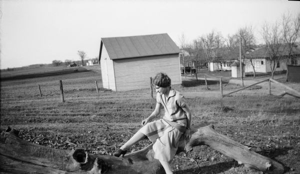 Mary Brandel posing on a weathered log, with fields, fence, and buildings in the background.
