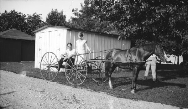A young man and woman (probably Mary Brandel) pose on a parked horse-drawn carriage next to a drive. A man (face obscured) is standing behind the horse on the right. There is a lake in the background, and two small buildings are on the left.