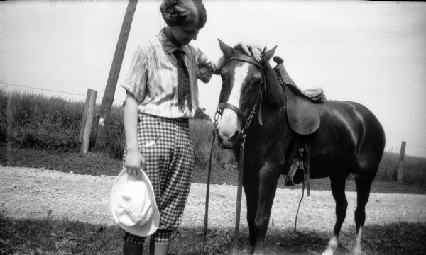 Mary Brandel poses in riding clothes with her pony. Behind them is a road, with a fence and field.