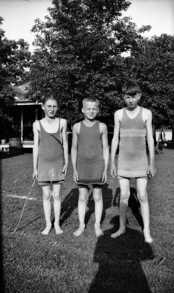 Three Taylor brothers pose in a line wearing bathing suits. Behind them is a cottage and a lake. The shadow of the photographer can be seen on the lawn.