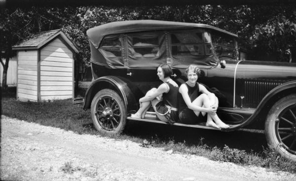 Mary Brandel and a friend pose on the sideboard of a parked car in what appear to be bathing suits. They are near a drive near a small building on the left and a cottage in the background.