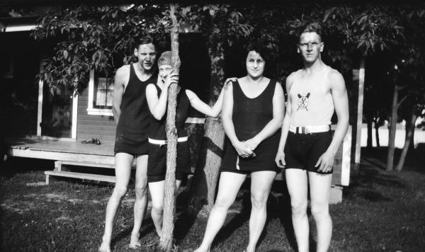 Mary Brandel, peeking from behind a tree, poses with two men and a woman. They are all wearing bathing suits. There is a cottage and a lake in the background.