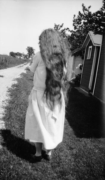 A view from behind of Mrs. Brandel, wife of E.W. Brandel, before she cut her long hair. She is outdoors near a road and small outbuildings.