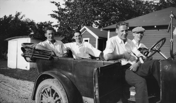 Four young men pose together in a car. They are parked on a drive near small outbuildings.