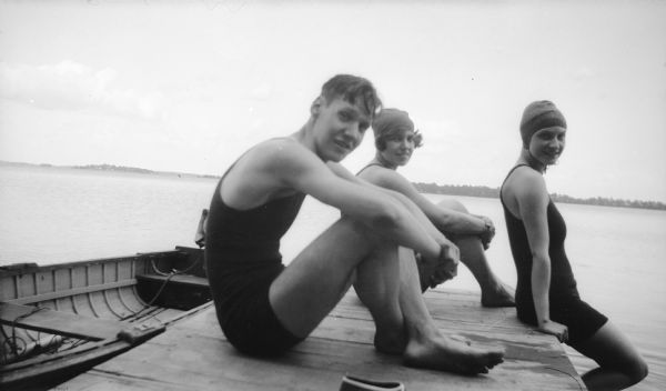 Mary Brandel, right, in bathing cap, poses with two friends on a pier to which a rowboat is moored. They are all wearing bathing suits. The far shoreline of the lake is in the background.