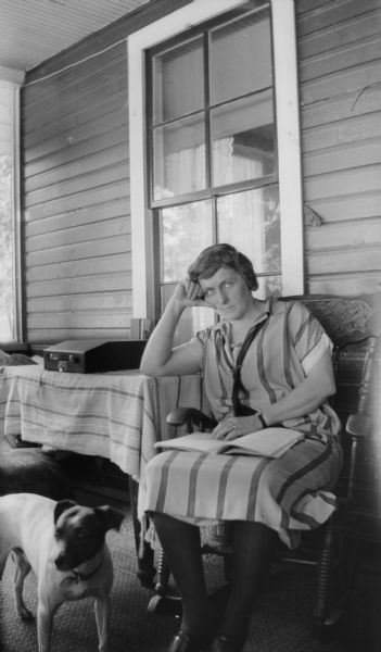 Mrs. E.W. Brandel sits in a rocking chair on the porch at Fox Lake leaning on the edge of a table. She has a book on her lap and Max the family dog is at her feet.