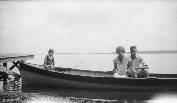 View from shoreline of young couple posing together in a boat by a pier. A doll is sitting on the bow of the boat. The far shoreline of the lake is in the background.