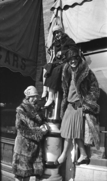 Three women pose together wearing fur coats and fashionable hats. The woman in the middle is perched on a column near a storefront, and the woman on the right is standing on a windowsill.