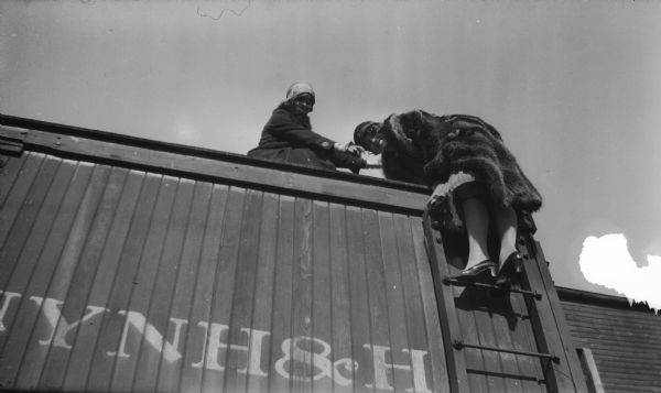 View looking up at two women in fashionable clothing and hats posing at the top of a railroad train car. One of the woman is standing on a ladder looking down.