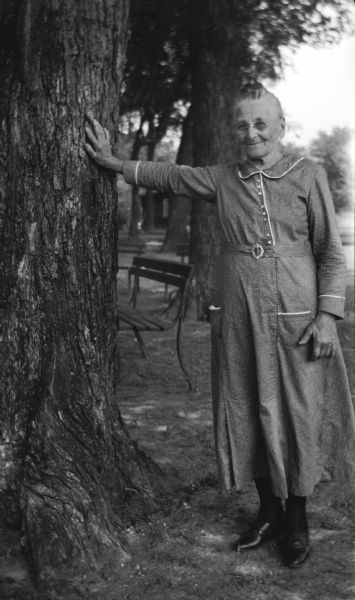 Aunt Lydia, a relative of the Brandel family, is wearing a dress and eyeglasses. She poses leaning against a tree.