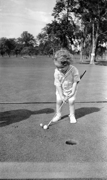 A toddler with curly hair plays golf. He is wearing a sailor shirt, shorts, and shoes.