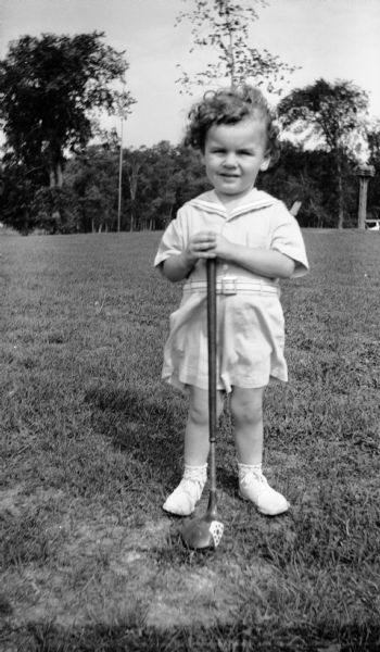 A little boy poses with a golf club. He is wearing a sailor shirt, shorts, and shoes. There is a birdhouse in the background.