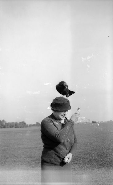 A woman stands on a golf course with a bird (a crow?)on her head. She is wearing a hat and sweater, and holds something (food?) in her right hand near the bird. She is holding a golf ball in her left hand.