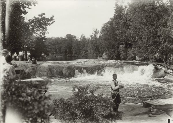 Group of people at Bear Trap Falls in the Wolf River near Keshena. People are standing on the shoreline on the left near a sign for the falls. A man is standing near a small footbridge on the right.