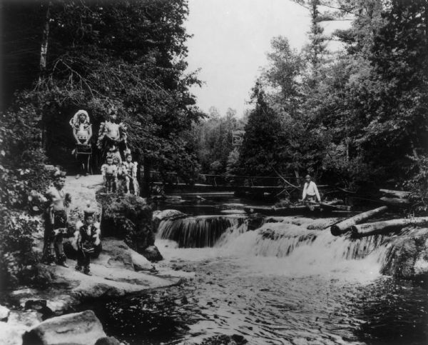 "Peavy Falls Group" of men and boys posed on bank of river. There is a man in a canoe in the river, and a footbridge in the background.