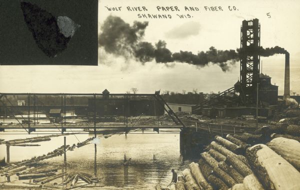 Stacks of logs are in the foreground with a man in a hat near the river. There is a chimney billowing smoke near the factory.