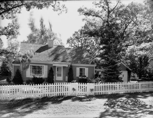 Exterior of the house of Byron and Mary Burch, 205 South Owen Drive. The yard is bordered by a picket fence and a car parks in the driveway on the right.