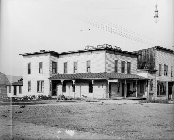 Exterior of Central Hotel on corner lot from street. There is a dog lying on the porch on the left, and a man relaxing in a chair with his feet on a post on the right.