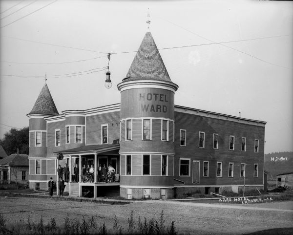 Exterior of Ward Hotel from road. Sign painted on roof of turret says, "Hotel Ward." A large group of men, two women, and a dog, are posing on the sidewalk and front porch of the hotel. A streetlight is suspended on wires above the road.