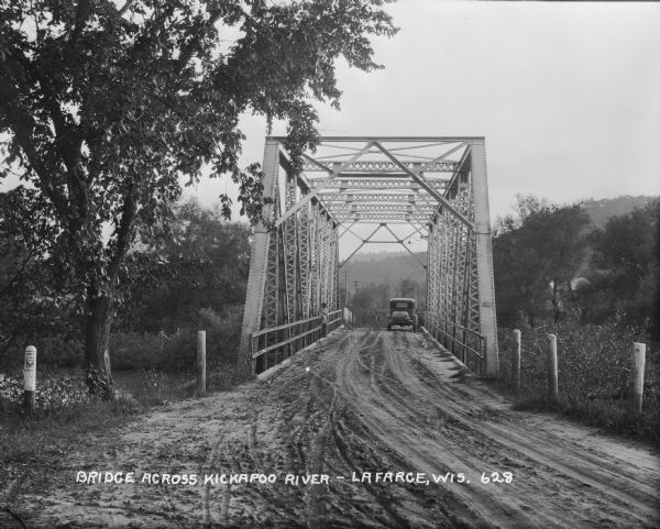 View down a metal bridge over the Kickapoo River showing a truck parked on the far side of the bridge. A man and a boy are standing at the rail of the bridge on the left. A post on the left is painted with a sign saying, "County Trunk M."