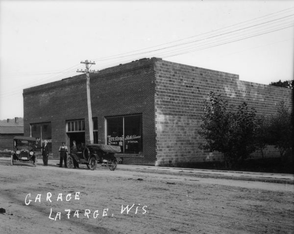 View from across street of four men posed next to two automobiles in front of a garage.