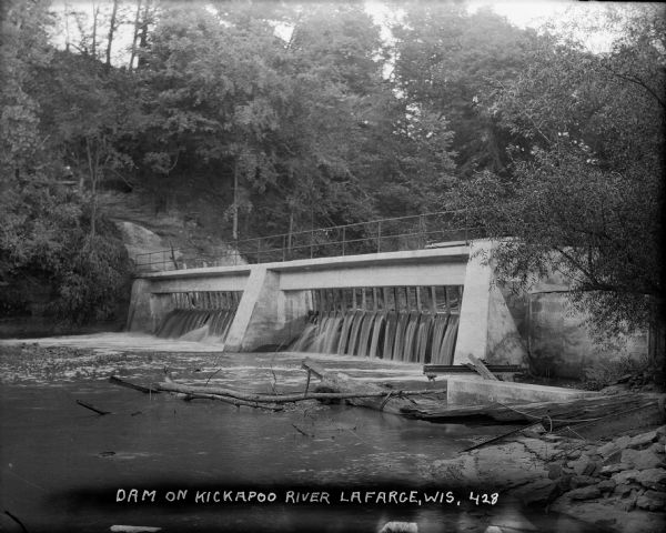 View from shoreline of dam on Kickapoo River. There is a steep hill on the left, and both sides of riverbank are covered with trees.