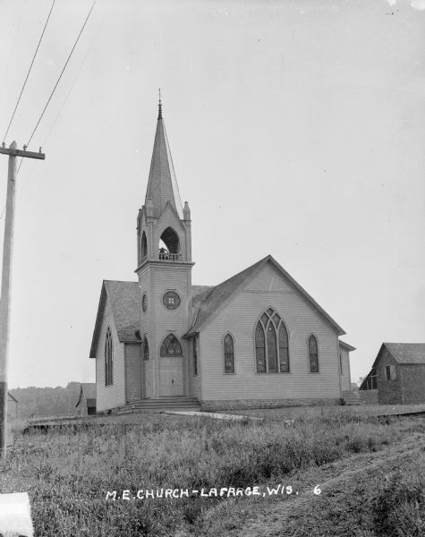 Exterior of Methodist Evangelical church. Small outbuildings are near church on the right. There is a bell in the belfry, and stained glass windows.