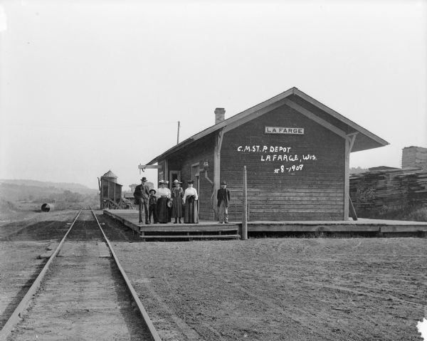 View from railroad tracks of a group of six people standing on the platform of the Chicago, Milwaukee and St. Paul depot. Beyond the depot is a water tower on a low platform. In the distance are low hills.