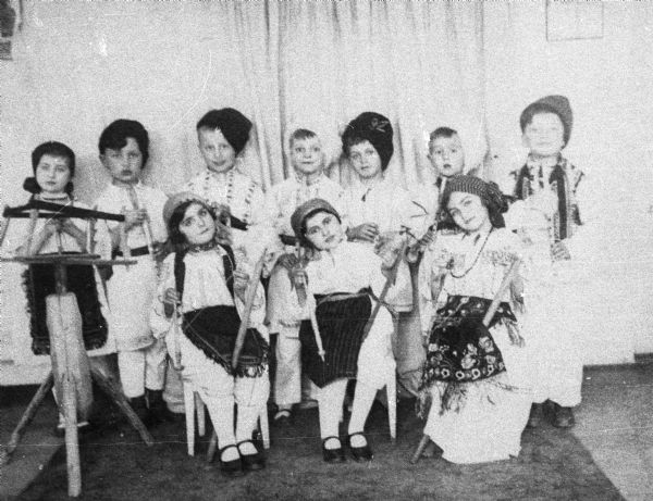 Magda Moses Herzberger (front row, far right) and her kindergarten class dressed in Romanian folk dress in Cluj, Romania. They are wearing caps or scarves on their heads, and are holding wooden spinning implements. There is a wooden yarn spinner on the left.