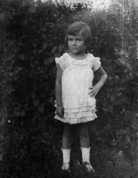 Magda Moses Herzberger standing in front of foliage in a light-colored dress with her left hand positioned on her hip.  Location identified as a resort in Avrig, Romania.