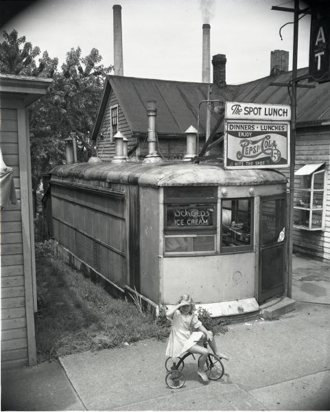 A little girl rides a tricycle in front of the Spot Lunch diner, a former streetcar. The Spot Lunch was located at 640 Williamson Street and owned by John B. Hanson.