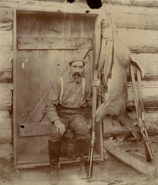 Man posed at door of a hunting lodge with guns, deer carcass, and two Northern pike.