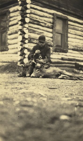 Man with a dog kneeling beside 2 deer carcasses in front of a log cabin.