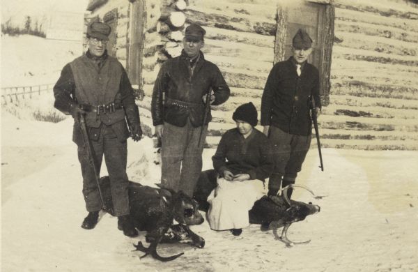 Three men standing holding guns with two deer carcasses on the ground. They are posed in front of a log cabin in the snow. A boy is seated on one of the deer carcasses. There is a For Rent sign on the side of the building on the left.