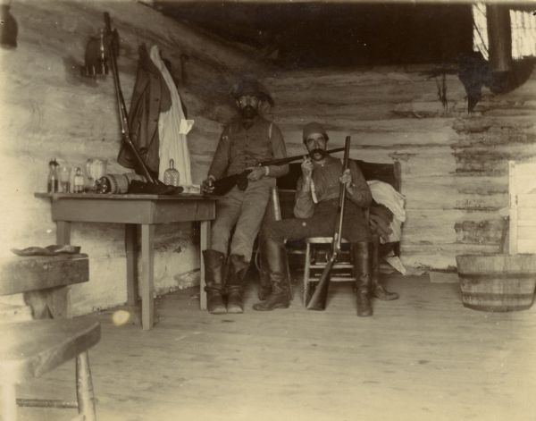 Two men posed with guns in a hunting cabin. The room in sparsely furnished with a table, chairs, and a bed.