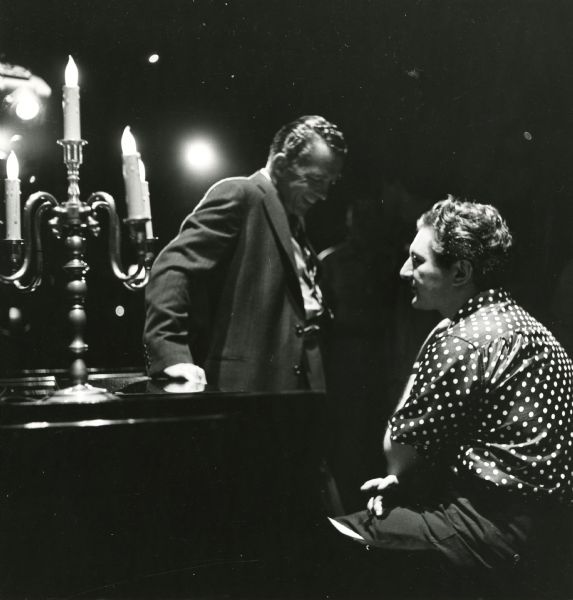 Ed Sullivan leans against a piano that Liberace is sitting near. There is a candelabra on the piano and stage lights are visible in the background.
