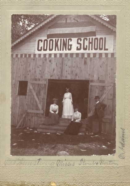 Misses Armstrong, Stout, and Norton, and Mister Adams posed on steps before the door of a building hosting a Farmers' Institute Cooking School.