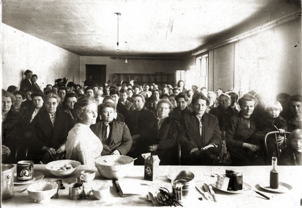 Room filled with women, some with children, attending a Farmers' Institute Cooking School class at Spring Valley, Wisconsin. There is a table with ingredients and utensils in the foreground.