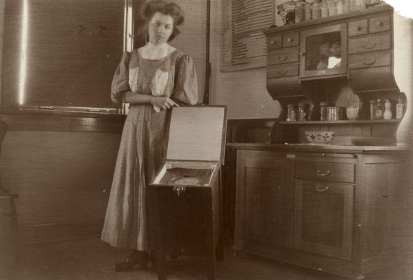 Woman in a long dress standing in kitchen with cook-box, a device to facilitate consistent results in cooking. There is a sideboard on the right.