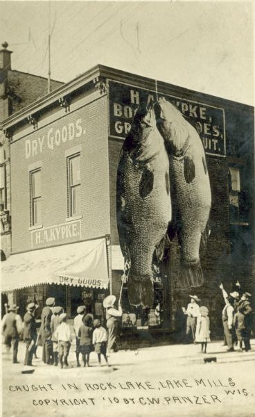 Photomontage of two largemouth bass hanging from the side of a building, with curious on-lookers observing the scene. The building is a dry goods store owned by H.A. Kypke. Caption reads: "Caught in Rock Lake, Lake Mills, Wis."
