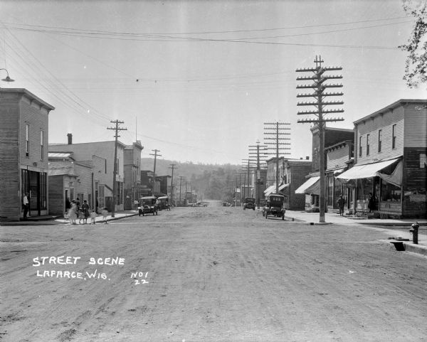 View down main street with storefronts on both sides, with large power lines along the sidewalks stretching into the distance, and a hill in the background. Cars parked near the curb. A group of women and children are on the left street corner, and a horse is in the distance.