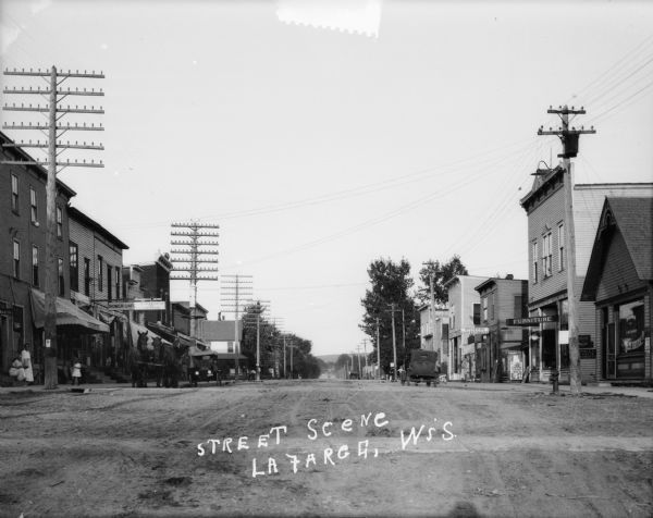 Street scene of road going through a business district with cars and horse-drawn vehicles parked along the curbs. Storefronts and power lines are along the road, and pedestrians are on the sidewalks.