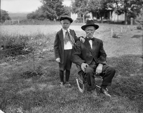 Outdoor portrait of man seated in a rocking chair with child standing next to him. They are both wearing hats and neckties. In the background is a house with a front porch.