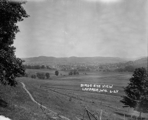 View over valley of town in the distance. A low hill is in the background. A fence and a path are near trees in the foreground.
