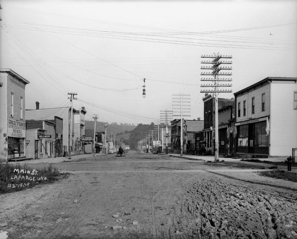 View down street showing commercial area with large power lines stretching into the distance, with a large hill in the background. A man is driving a horse-drawn vehicle down the dirt road near storefronts.
